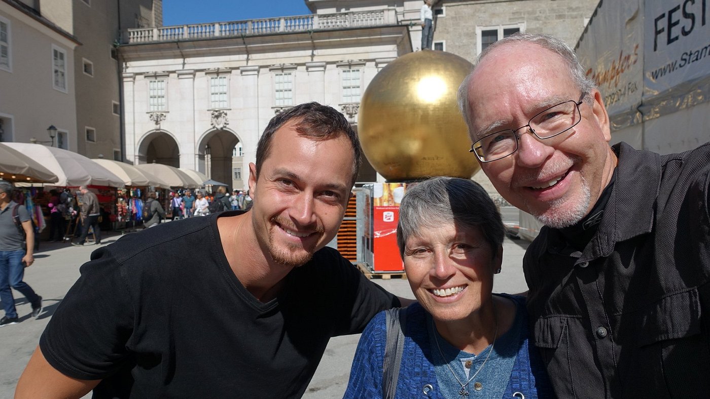 Drei Personen lächeln für ein Selfie im Freien auf einem Stadtplatz, mit einer großen goldenen Kugel und Gebäuden im Hintergrund.