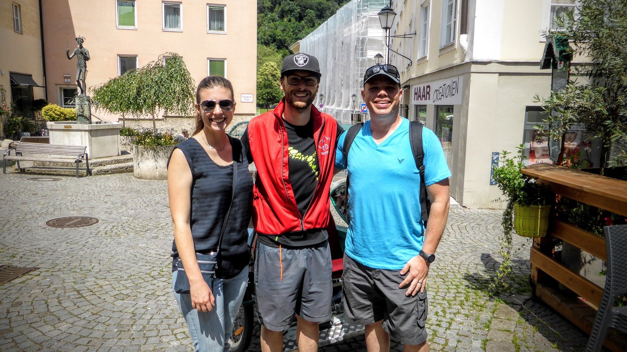Drei Personen stehen auf einer Kopfsteinpflasterstraße in einer europäischen Stadt, mit Gebäuden und Grünflächen im Hintergrund, und lächeln in die Kamera.