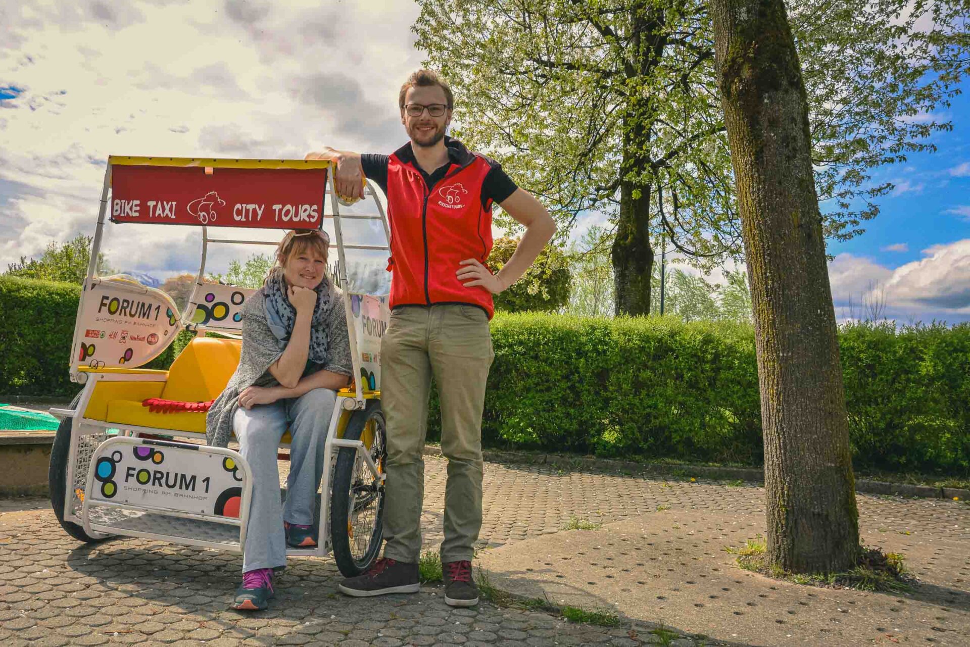 Zwei Personen stehen neben einem Fahrradtaxi mit der Aufschrift "City Tours" in einer Parkanlage mit Bäumen und einem bewölkten Himmel im Hintergrund.
