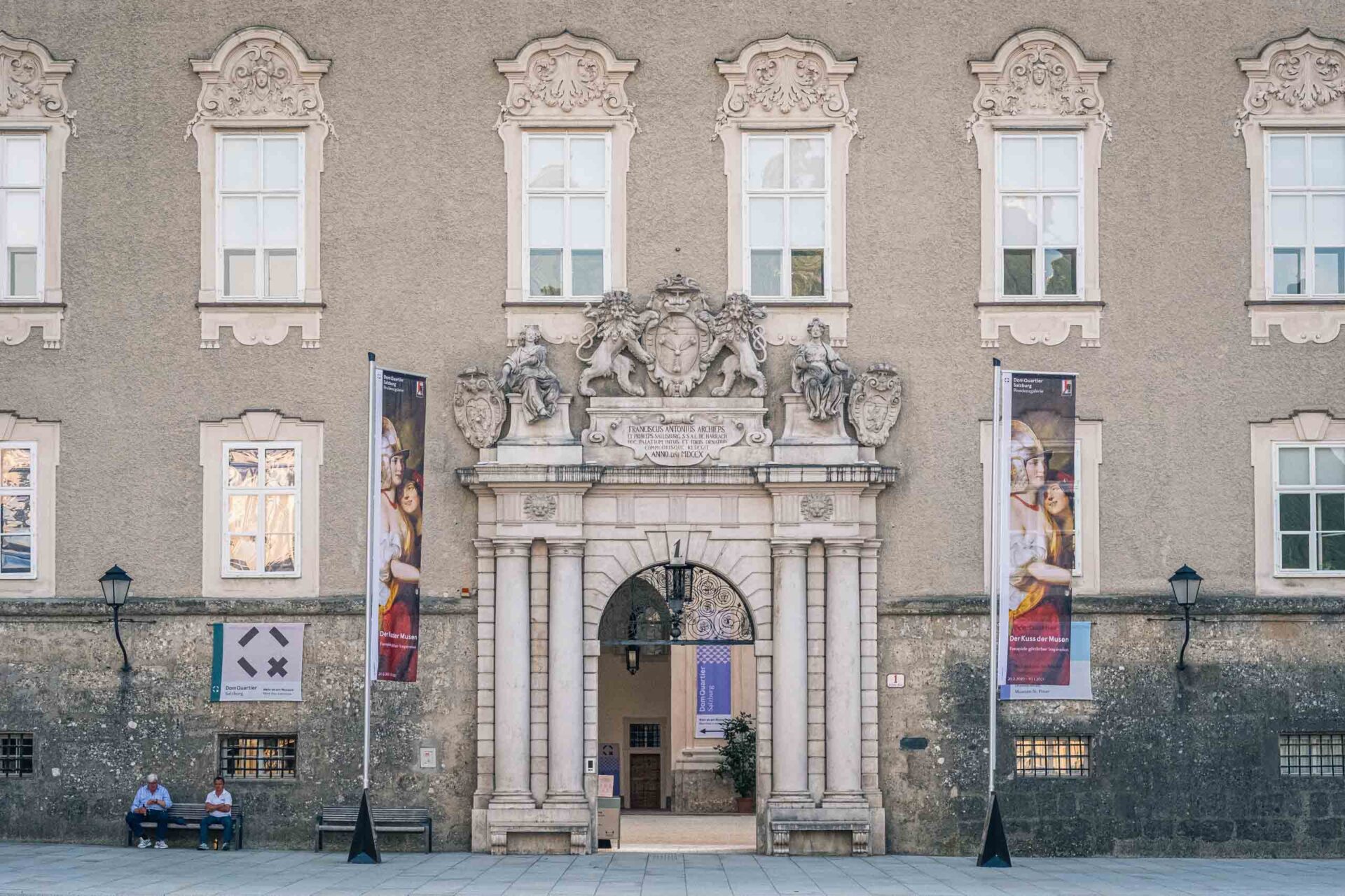 Verschnörkelte Steinfassade mit bogenförmigem Eingang, dekorativen Skulpturen über der Tür, zwei Bannern auf beiden Seiten und zwei Personen, die links auf einer Bank sitzen.