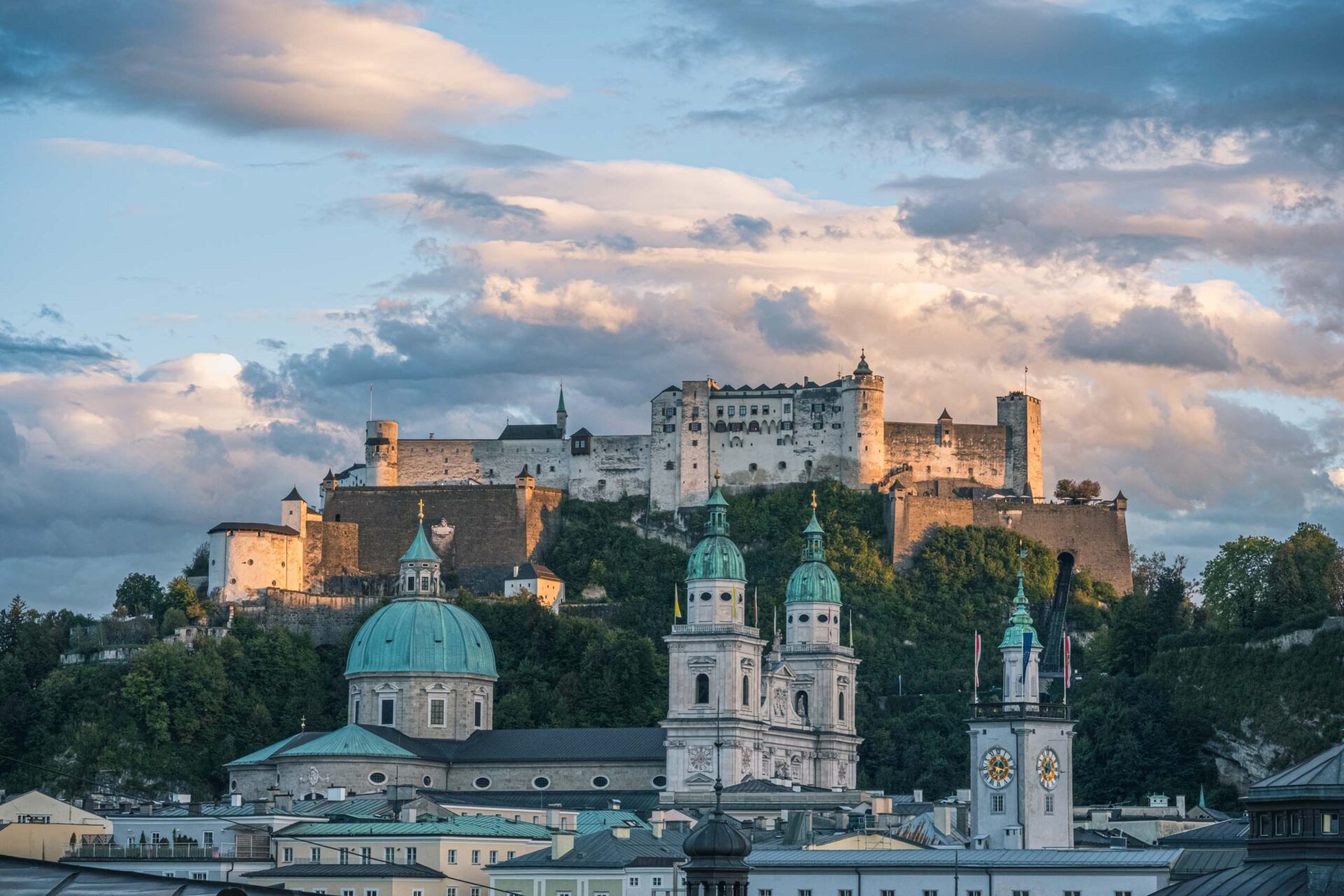 Eine große steinerne Festung thront auf einem Hügel unter einem wolkenverhangenen Himmel, während in der Stadt darunter Kirchenkuppeln und Uhrentürme zu sehen sind.