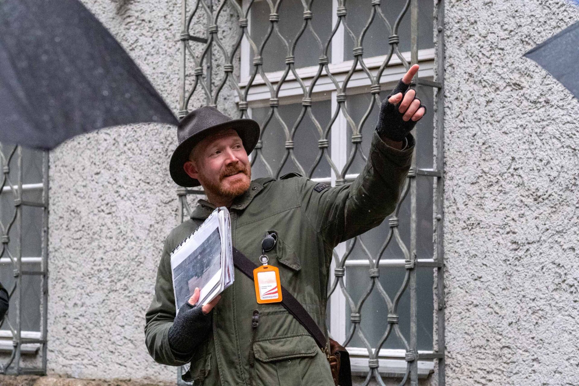 Ein Mann in grünem Mantel und Hut hält Papiere in der Hand und gestikuliert, während er draußen vor einem vergitterten Fenster spricht, während Regenschirme einen Teil des Bildes verdecken.