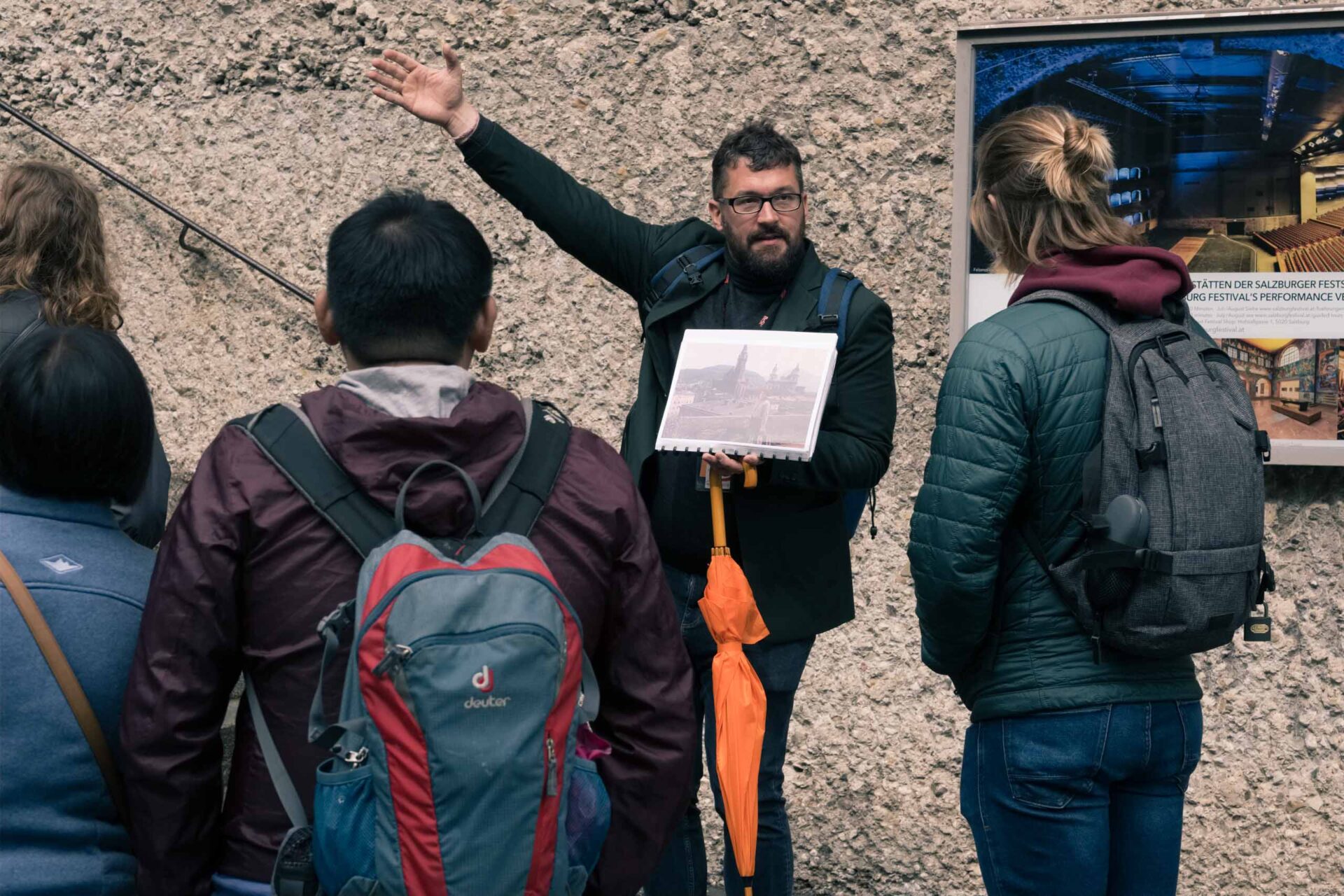 Ein Mann mit einem Foto und einem orangefarbenen Regenschirm führt eine Gruppe auf einem Rundgang und deutet auf eine strukturierte Wand mit einem Informationsplakat.