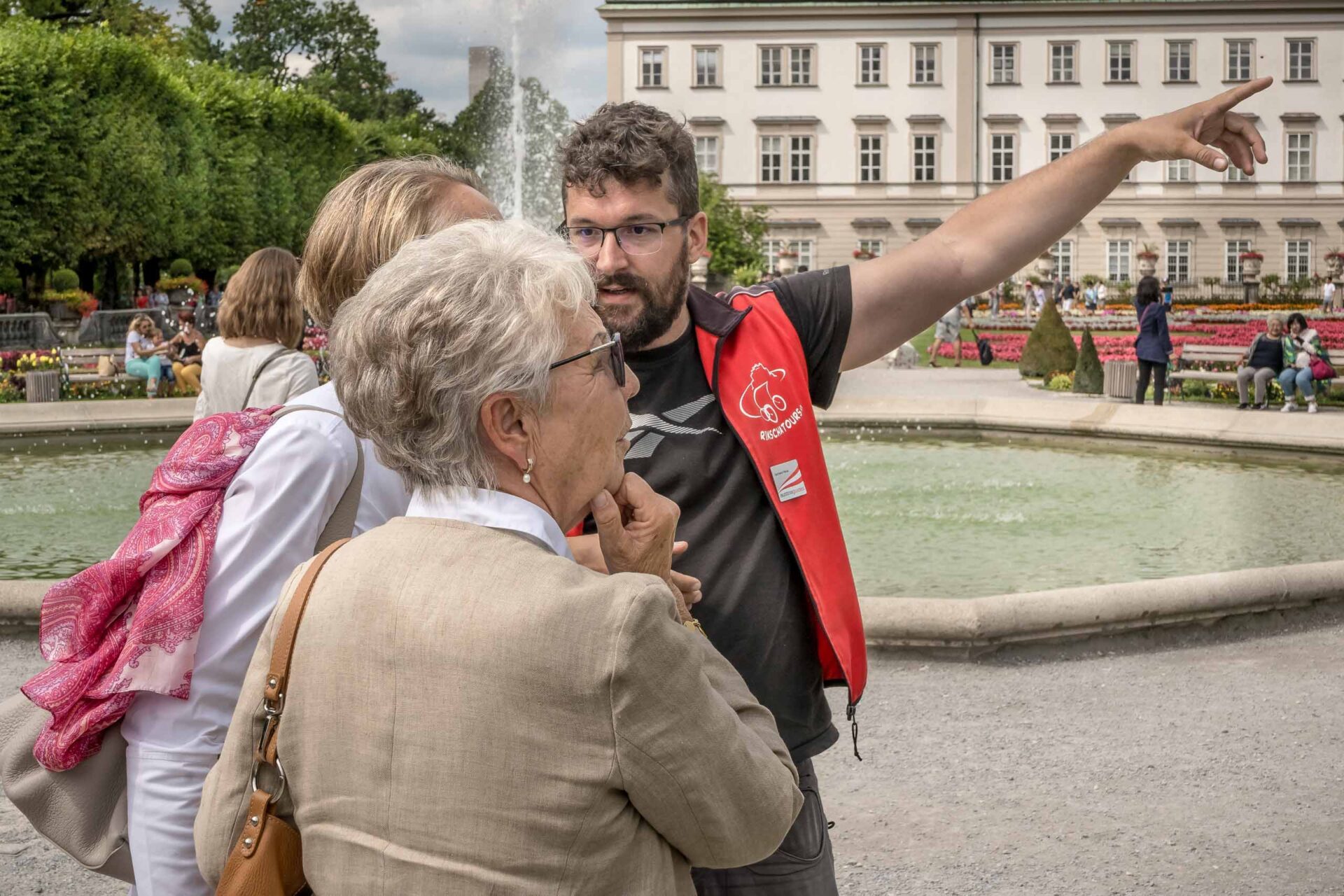 Ein Mann in roter Weste gestikuliert und spricht mit zwei Frauen in der Nähe eines Springbrunnens in einem Landschaftspark, mit einem offiziellen Gebäude im Hintergrund.
