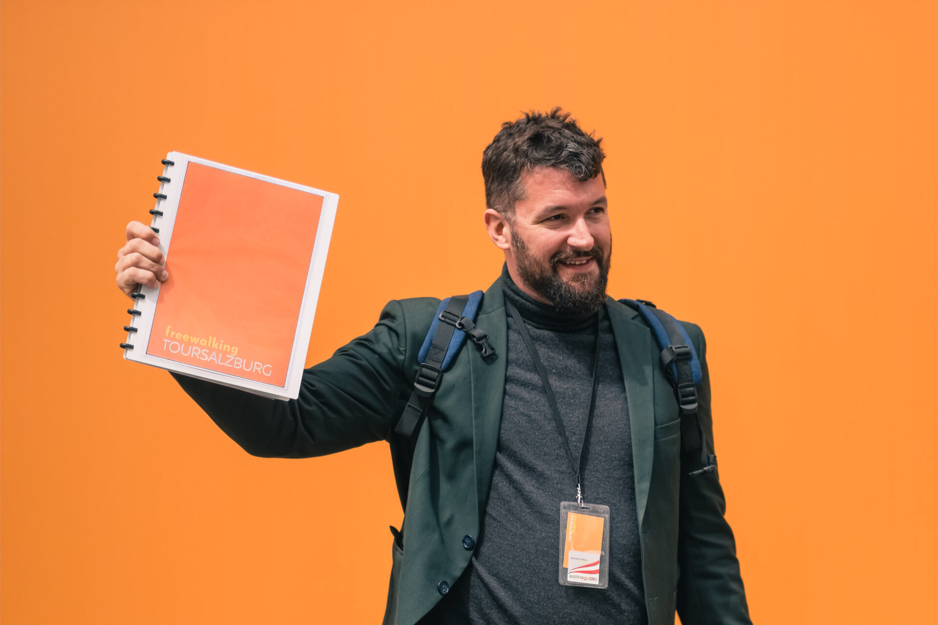 Ein Mann in einem dunklen Blazer hält eine orangefarbene Mappe mit der Aufschrift "TOUR SALZBURG" vor einem orangefarbenen Hintergrund hoch. Er trägt einen Rucksack und ein Schlüsselband.