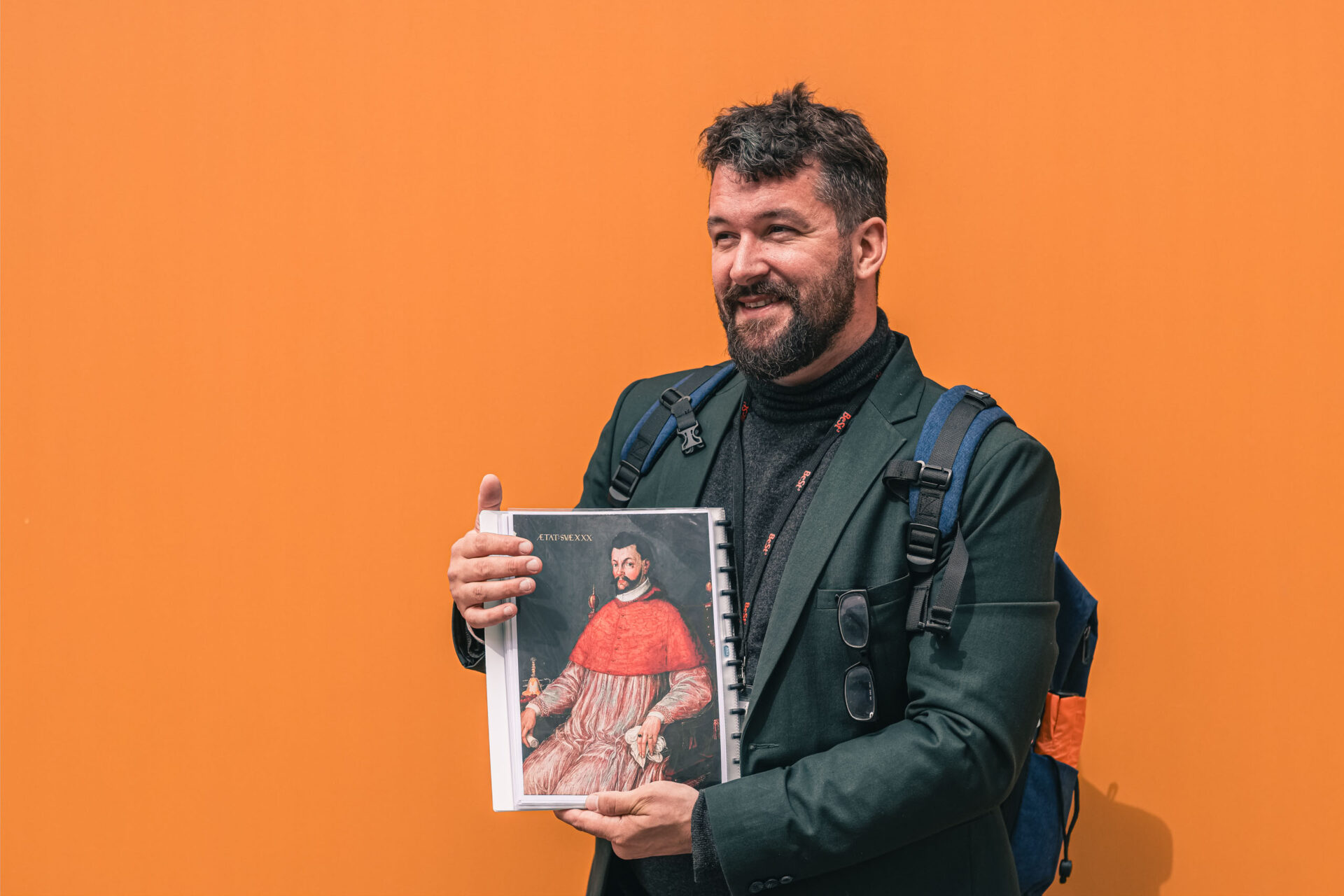 Ein Mann mit einem Rucksack steht an einer orangefarbenen Wand und hält ein Buch hoch, auf dessen Einband das Porträt eines Mannes in einer roten Robe abgebildet ist.