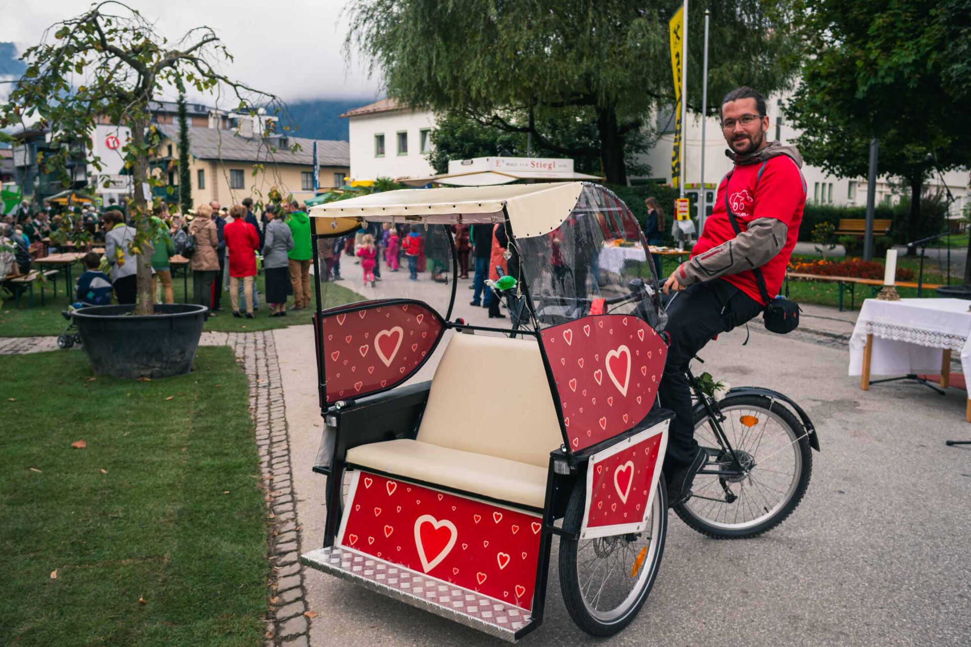 Ein Mann in einem roten Hemd lächelt, während er auf einer dekorierten Rikscha mit Herzmotiven bei einer Veranstaltung im Freien sitzt. Im Hintergrund versammeln sich Menschen an Tischen und Bäumen.