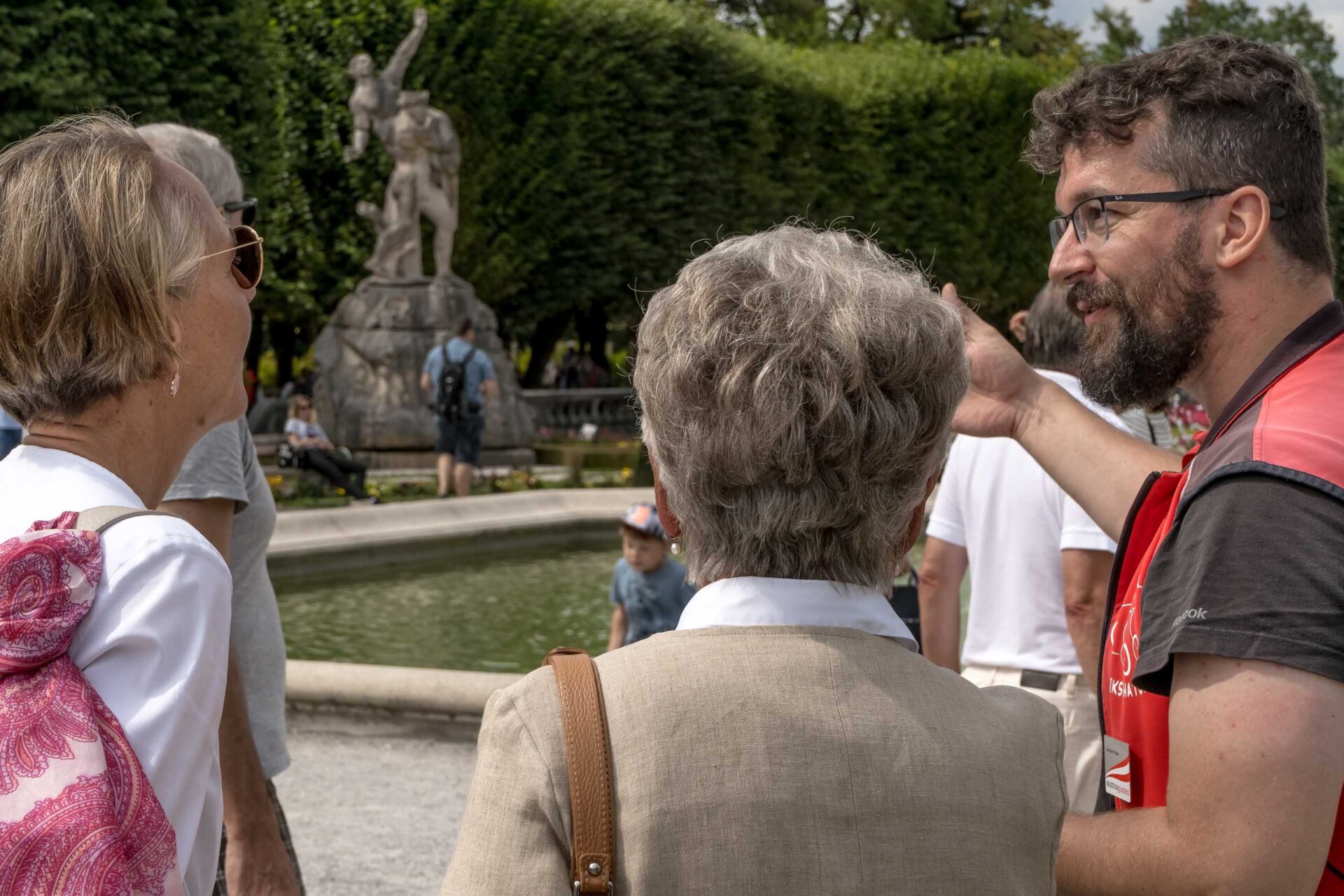 Ein Fremdenführer gestikuliert, während er mit zwei Frauen in der Nähe eines Brunnens mit einer Statue in einem Park spricht, umgeben von anderen Besuchern.