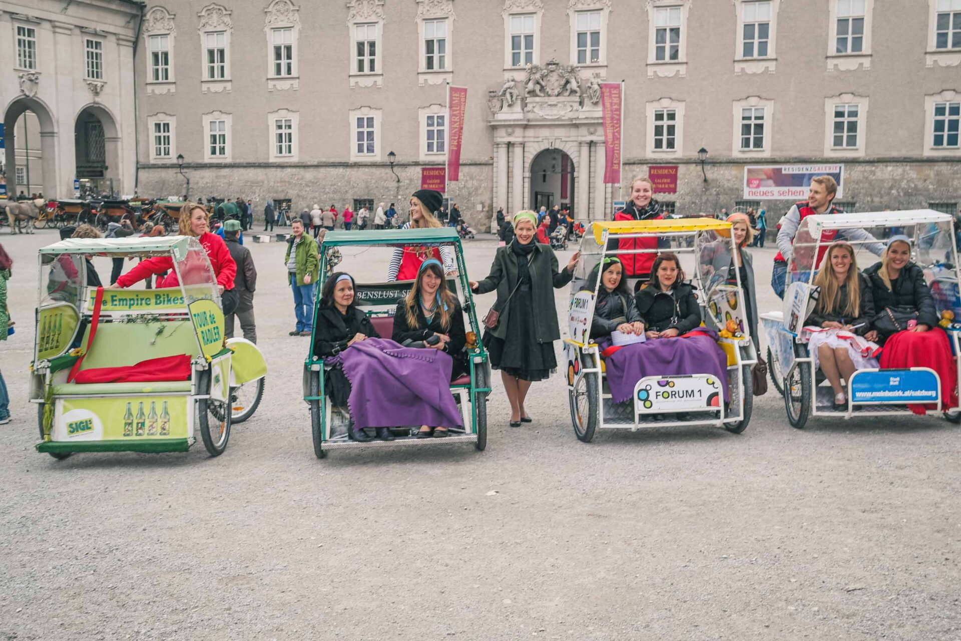 Eine Gruppe von Menschen sitzt in dekorierten Tretautos, die auf einem Platz geparkt sind, mit einem historischen Gebäude im Hintergrund.
