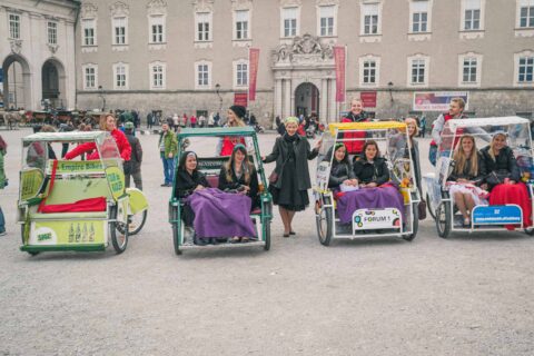 Eine Gruppe von Menschen sitzt in dekorierten Tretautos, die auf einem Platz geparkt sind, mit einem historischen Gebäude im Hintergrund.