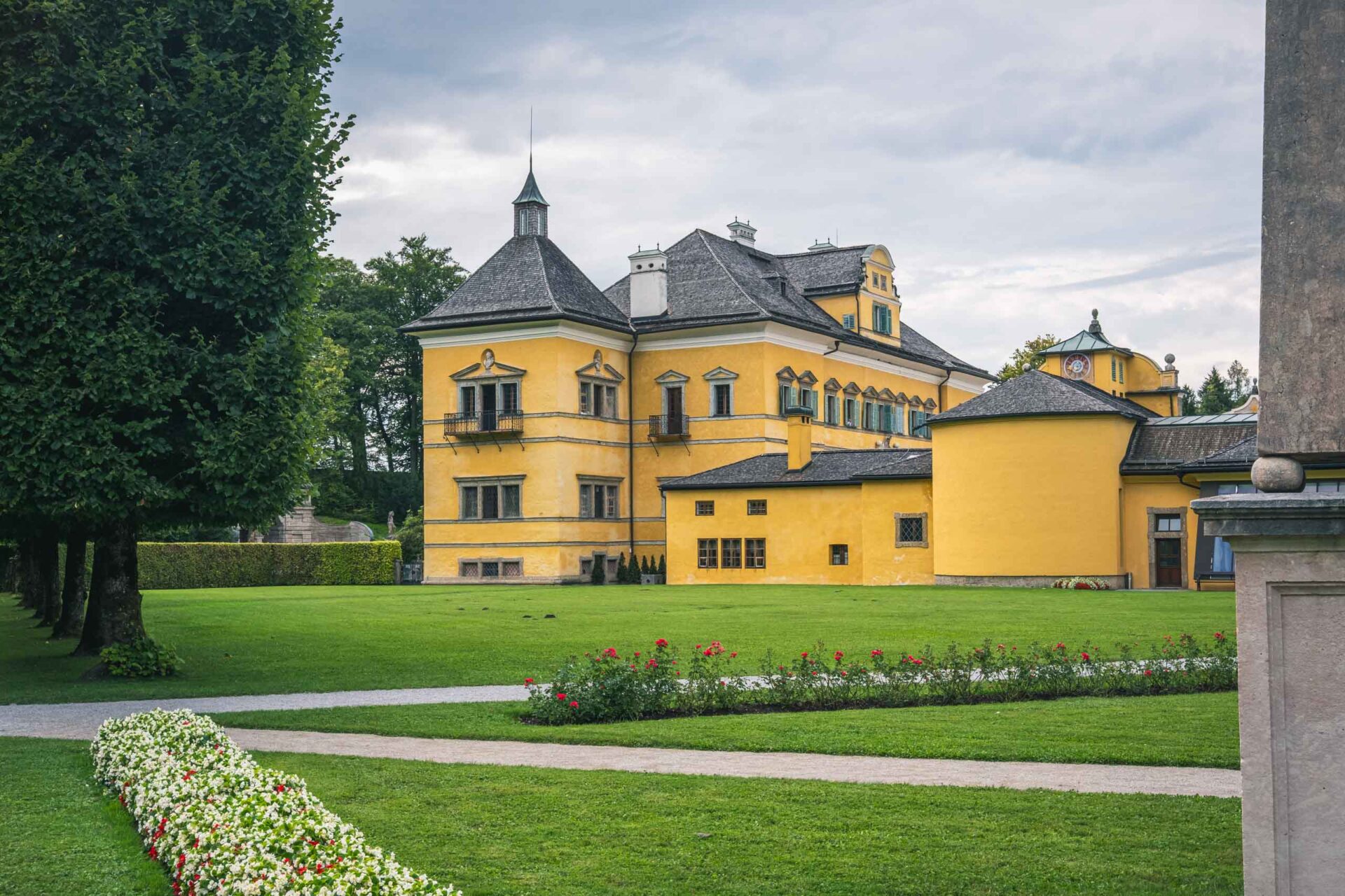 Ein großes gelbes historisches Gebäude mit mehreren Dächern und Türmen steht auf einem gepflegten Rasen mit Blumenbeeten und gepflasterten Gehwegen, die von Bäumen gesäumt sind.