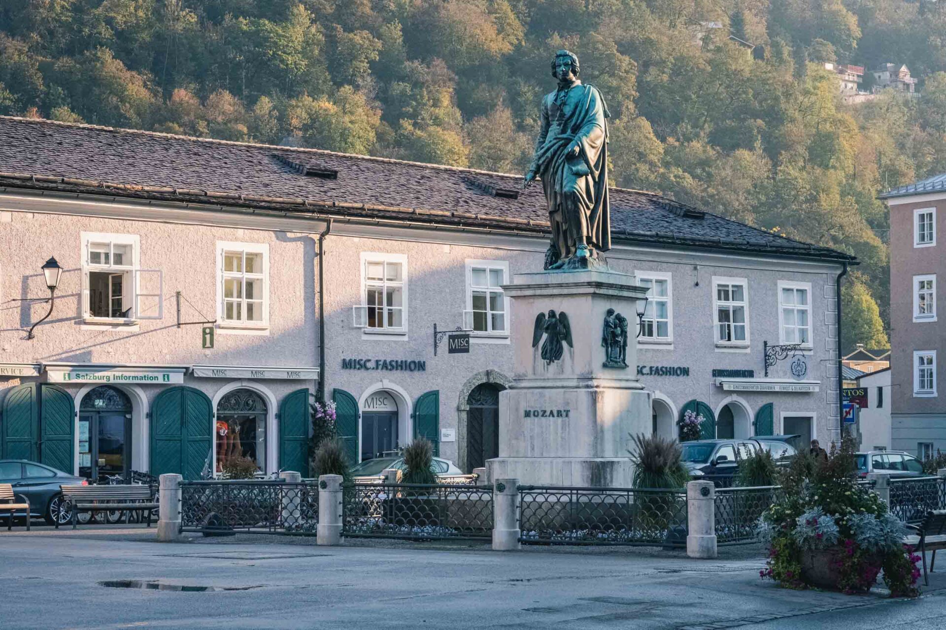 Eine Bronzestatue von Mozart steht auf einem Sockel auf einem Stadtplatz, mit Geschäften und Autos im Hintergrund und bewaldeten Hügeln dahinter.