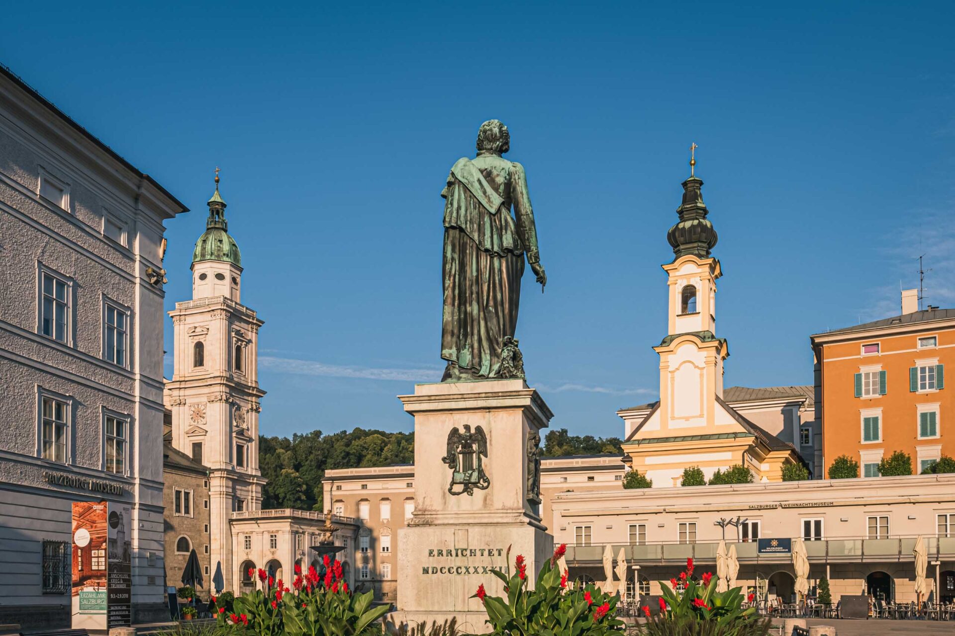 Bronzestatue einer stehenden Figur auf einem Sockel auf einem Stadtplatz, umgeben von historischen Gebäuden und blühenden Pflanzen unter einem klaren blauen Himmel.