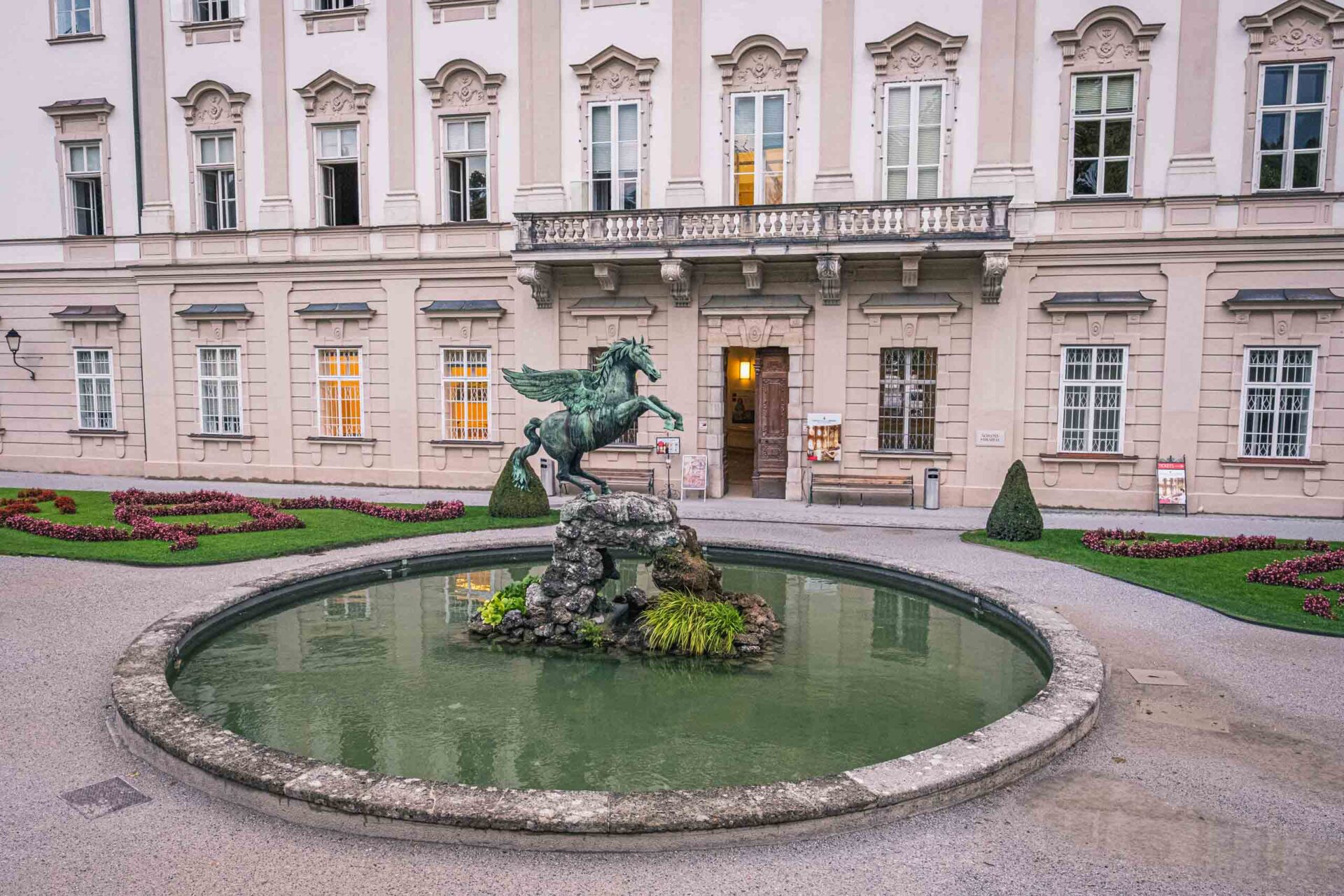 Eine bronzene Pegasus-Statue steht auf Felsen in der Mitte eines runden Brunnens vor einem großen historischen Gebäude mit Blumenbeeten in der Nähe.