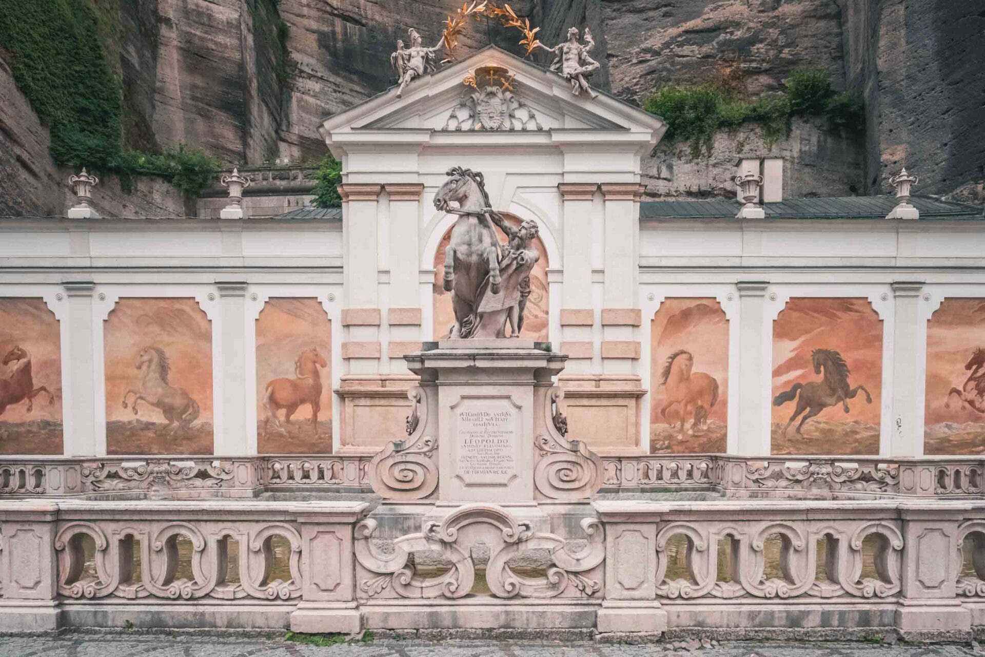 Vor einer Wand mit gemalten Pferdemalereien steht ein steinerner Pferdebrunnen, der von einem kunstvollen Geländer umgeben ist und sich an eine felsige Klippe schmiegt.