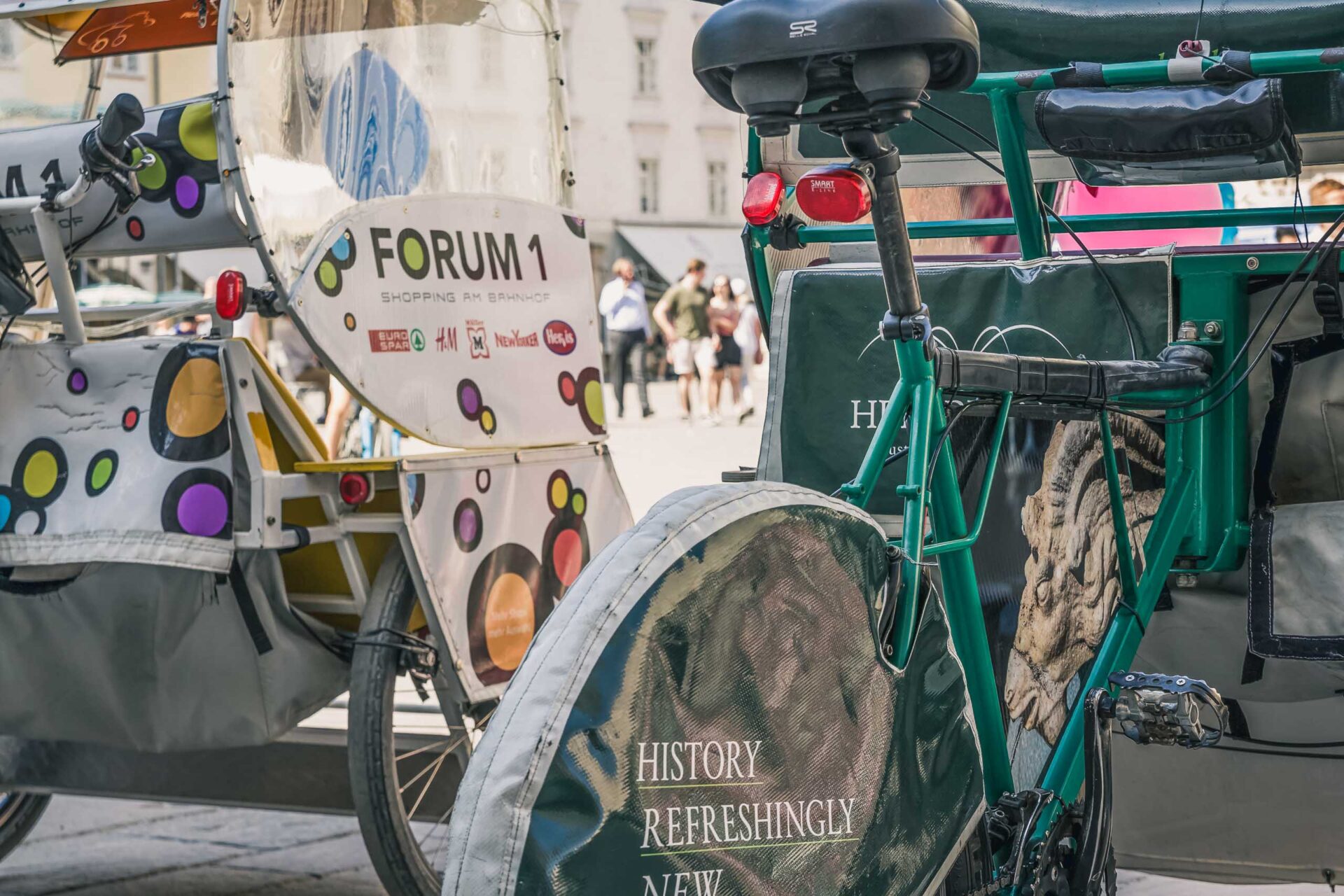 Nahaufnahme von zwei geparkten Fahrradtaxis mit Werbedecken und bunten Motiven auf einer Stadtstraße, mit Menschen im Hintergrund.