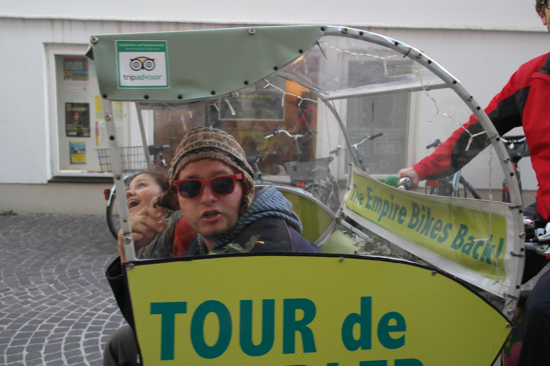 Zwei Personen sitzen in einem Fahrradtaxi mit "Tour de"-Schild und TripAdvisor-Logo, die eine trägt eine rote Sonnenbrille und einen Hut, die andere lächelt und zeigt in die Kamera.