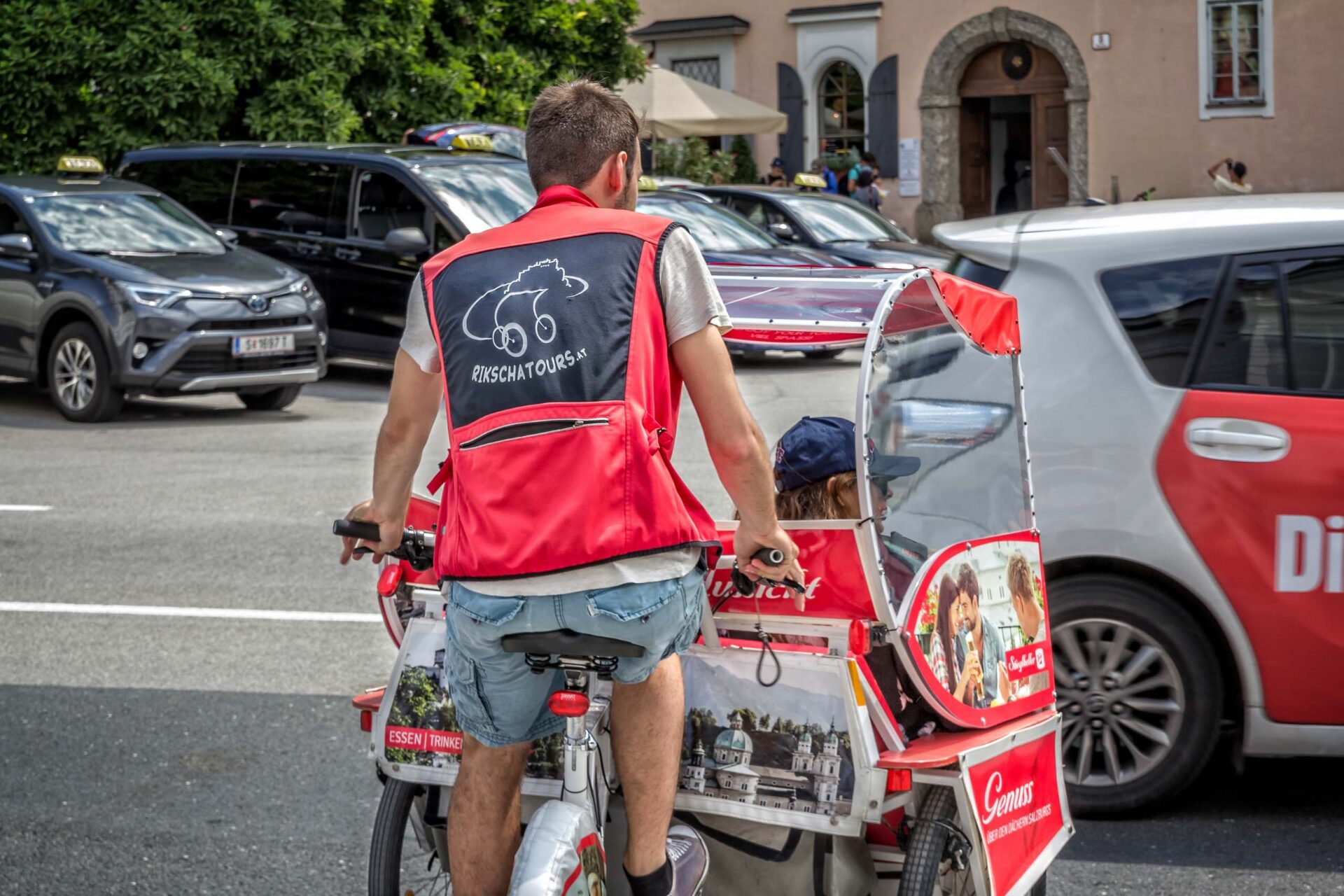 Eine Person mit roter Weste fährt mit einem Fahrgast in einem Fahrradtaxi in einem städtischen Gebiet mit Autos und Gebäuden im Hintergrund.