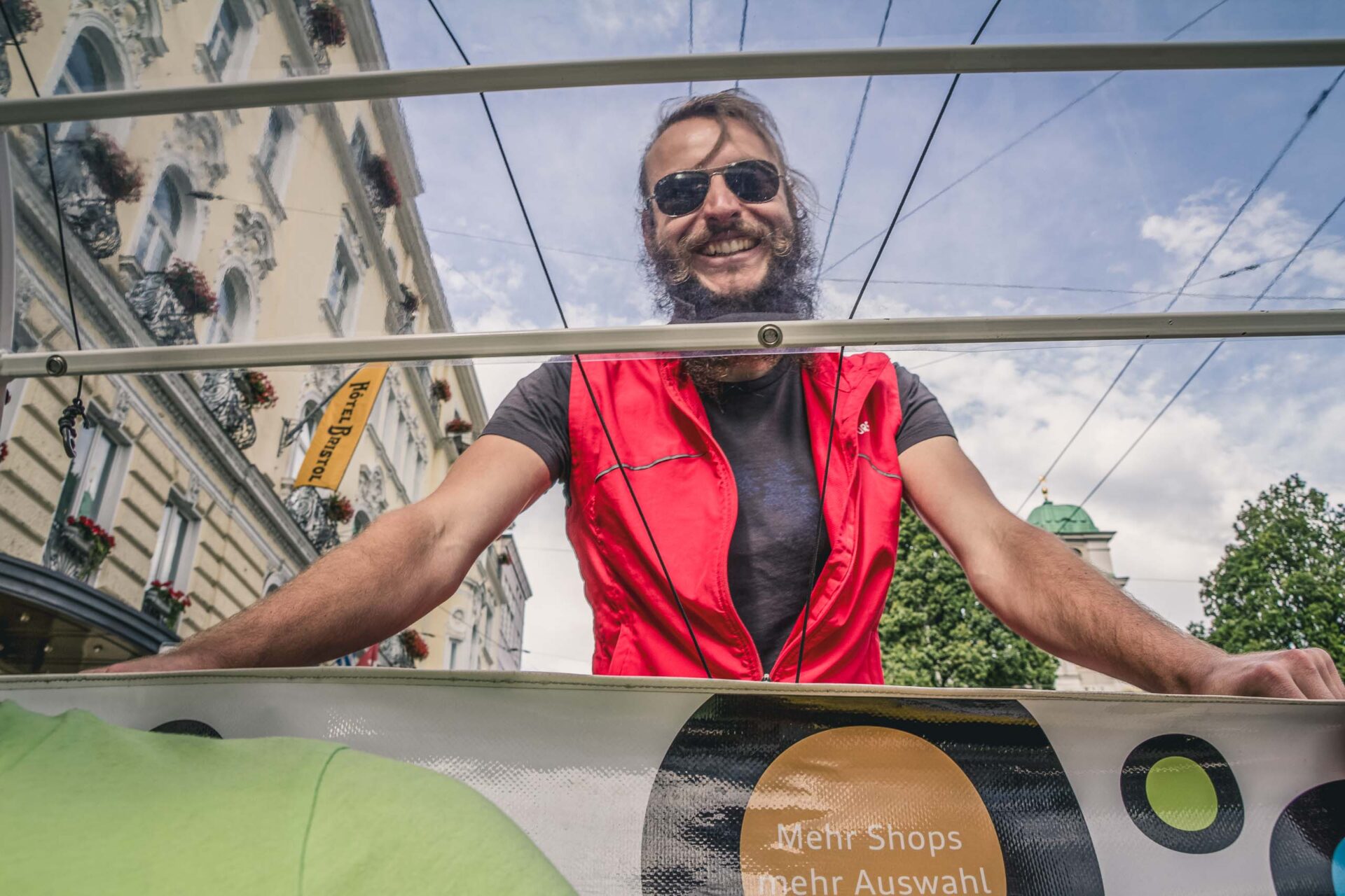 Ein Mann mit Bart und Sonnenbrille, der eine rote Weste trägt, lächelt, während er im Freien in der Nähe eines Straßenbahn- oder Buswartehäuschens in einer städtischen Umgebung steht.