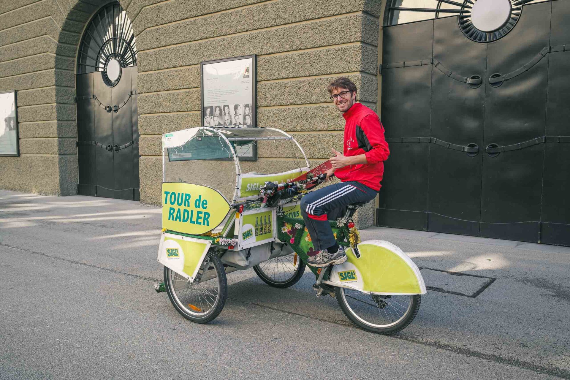 Eine Person in einer roten Jacke sitzt in einem gelb-weißen Fahrradtaxi mit der Aufschrift "Tour de Radler", das auf einer Straße in der Stadt geparkt ist.