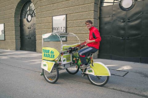 Eine Person in einer roten Jacke sitzt in einem gelb-weißen Fahrradtaxi mit der Aufschrift "Tour de Radler", das auf einer Straße in der Stadt geparkt ist.