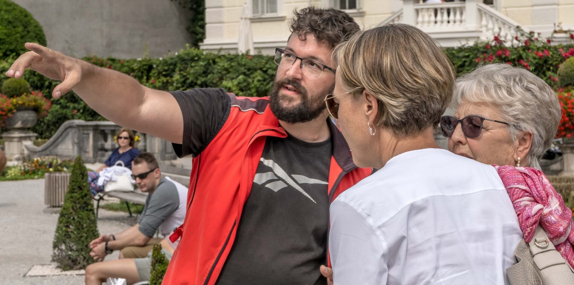 Ein Mann in einer roten Weste gestikuliert und zeigt auf etwas, während er sich mit zwei Frauen in einem Garten unterhält.