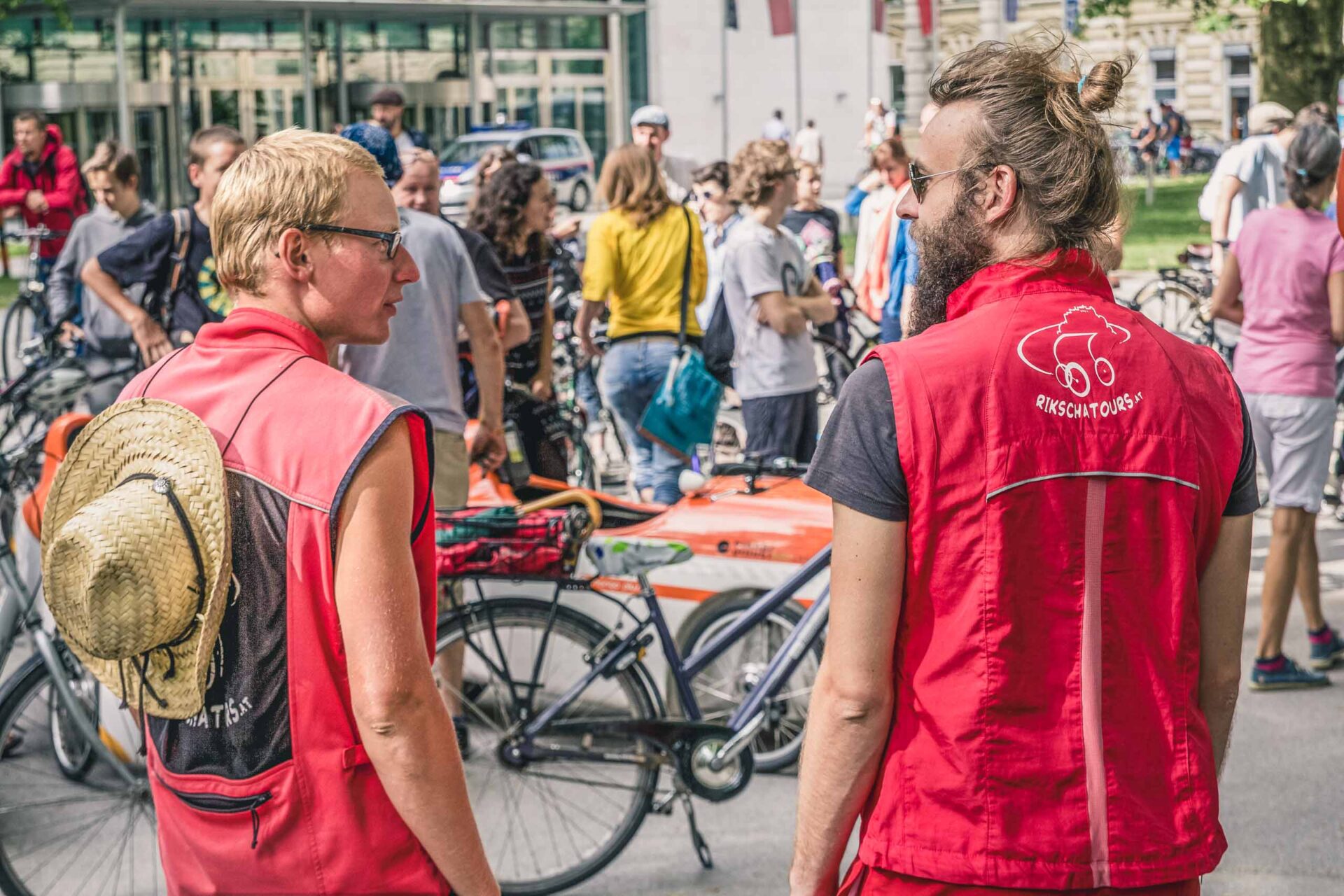 Zwei Personen in roten Westen stehen in der Nähe von Fahrrädern und blicken in eine Menschenmenge im Freien. Eine hat einen Strohhut auf dem Rücken; beide haben kurze Haare. Im Hintergrund sind weitere Menschen und Fahrräder zu sehen.