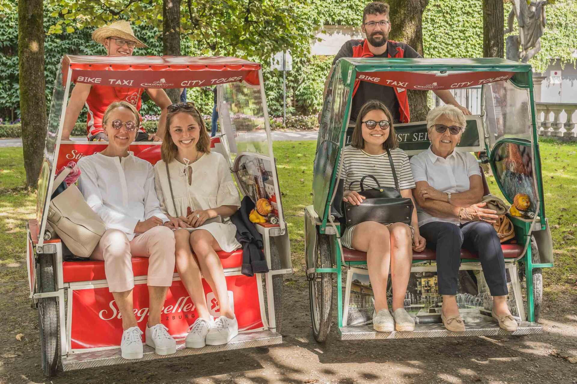 Zwei Frauenpaare sitzen in getrennten Rikschas, die nebeneinander in einem schattigen Park geparkt sind, wobei hinter jeder Rikscha ein Mann steht.