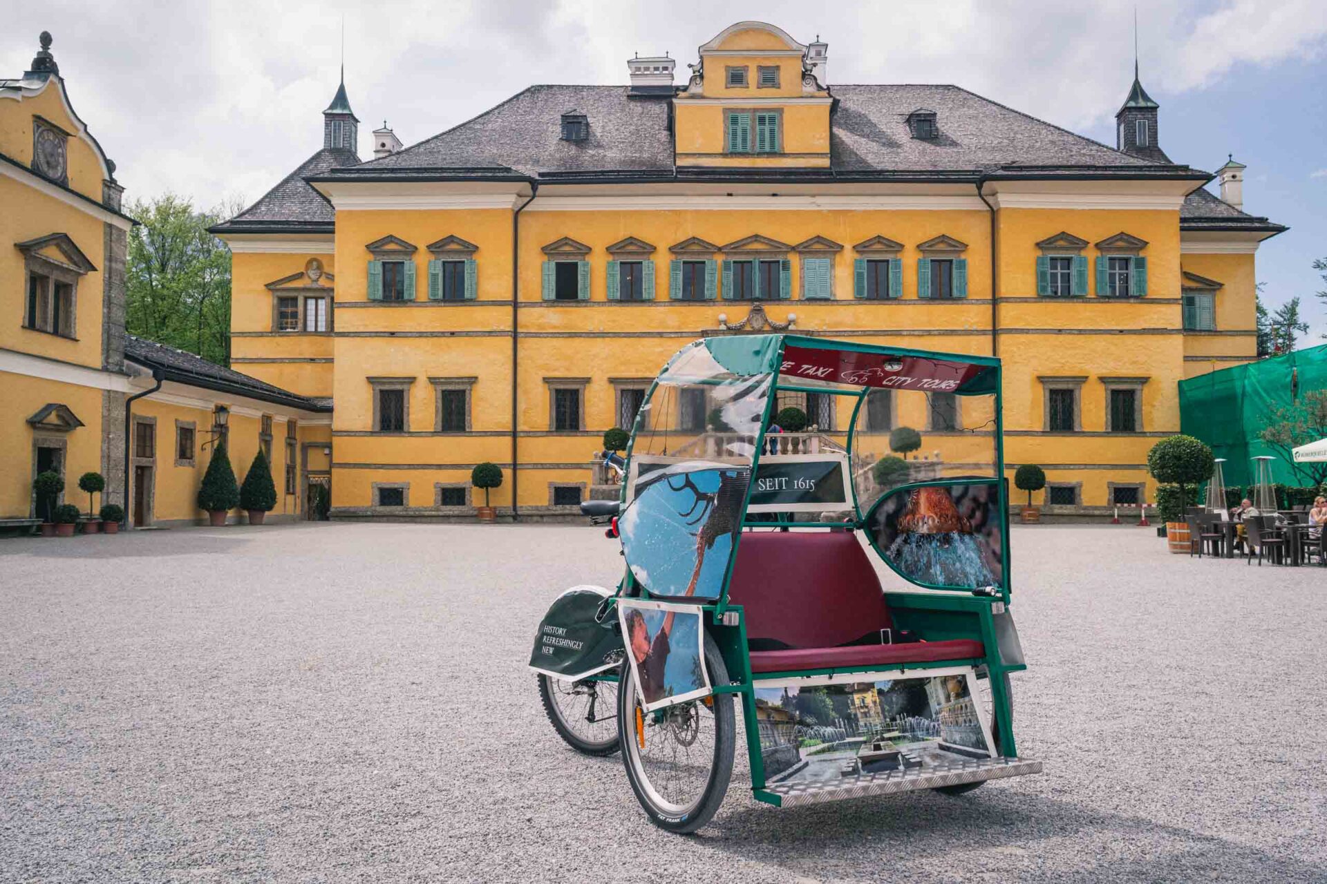 Eine bunte Rikscha parkt vor einem großen, gelben historischen Gebäude mit vielen Fenstern und einem Hof aus Kieselsteinen.