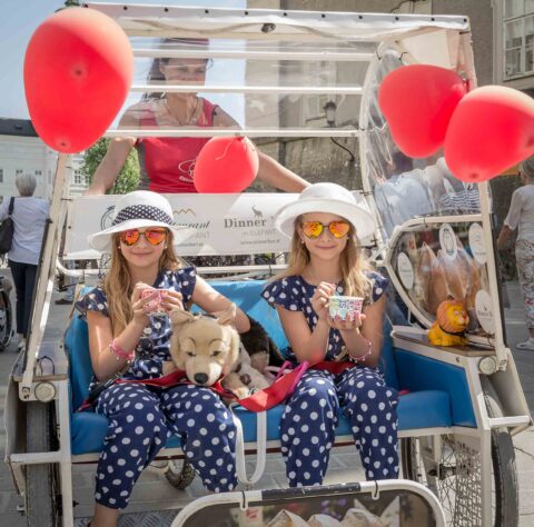 Zwei Mädchen in passenden Polka-Dot-Kostümen und Hüten sitzen in einer geschmückten Rikscha und halten Getränke in der Hand, während eine Frau am Steuer sitzt und ein ausgestopfter Hund neben ihnen liegt. Rote Luftballons sind an der Rikscha befestigt.
