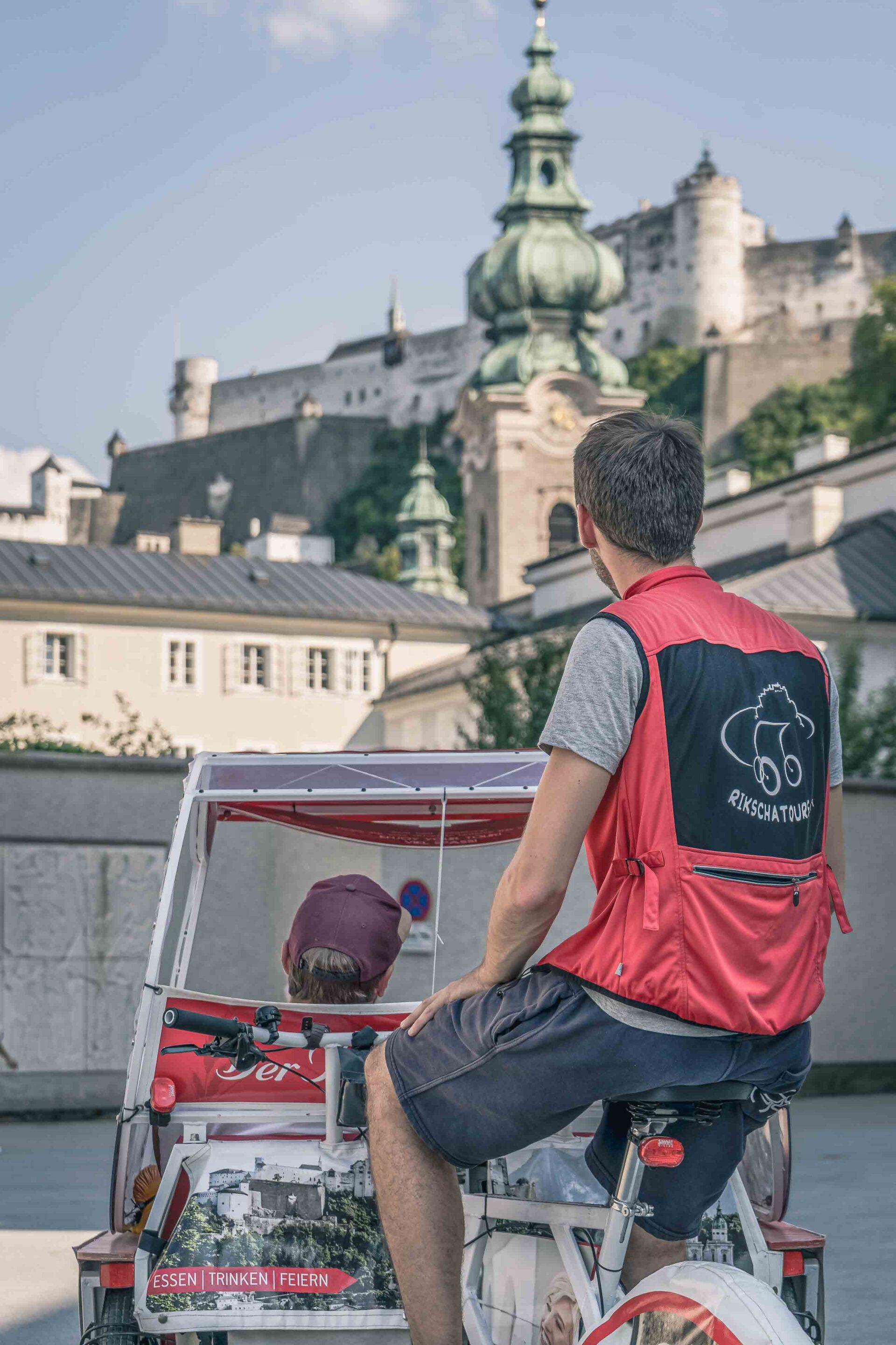 Ein Mann in roter Weste fährt mit einer Rikscha und einem Fahrgast durch eine Stadt mit historischen Gebäuden und einer Burg im Hintergrund.