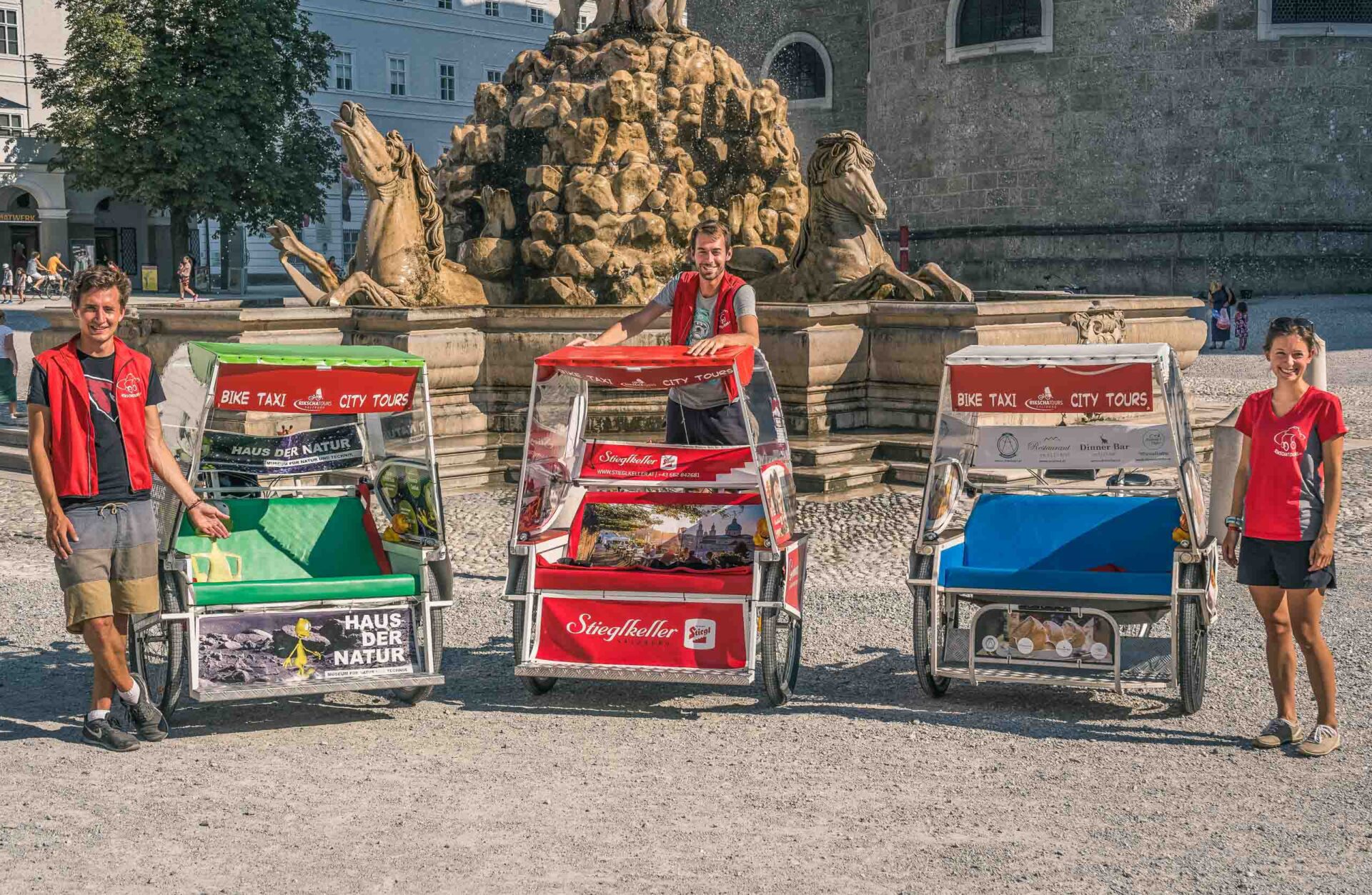 Vier Personen stehen im Freien neben drei bunten Fahrrad-Rikschas mit Werbeschildern, in der Nähe eines Steinbrunnens mit Pferdeskulpturen im Hintergrund.
