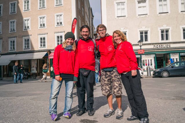Vier Personen in passenden roten Jacken stehen zusammen und lächeln in die Kamera auf einem europäischen Stadtplatz mit Gebäuden und ein paar Fußgängern im Hintergrund.