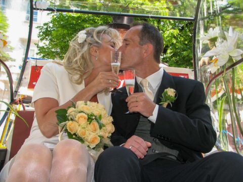 Braut und Bräutigam küssen sich mit Sektgläsern und einem Strauß gelber Rosen in der Hand, während sie an ihrem Hochzeitstag im Freien sitzen.