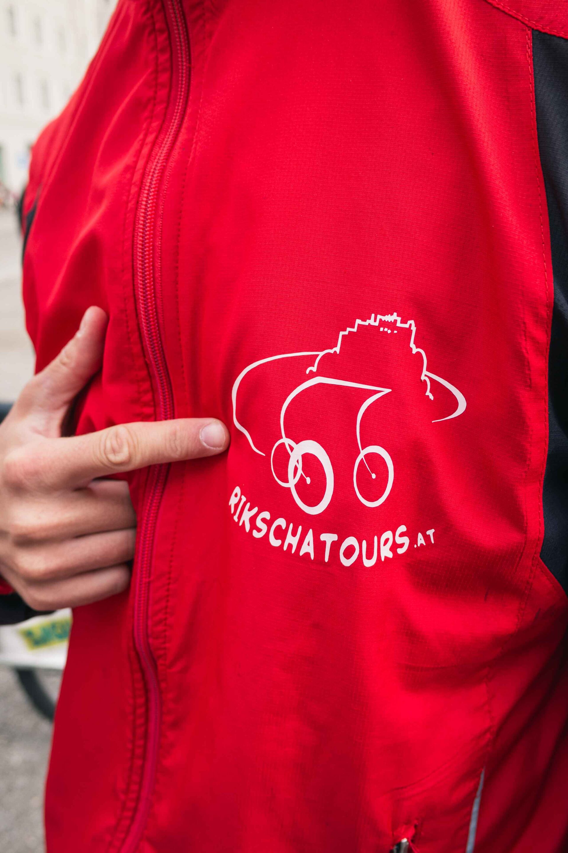 Eine Person zeigt auf ein weißes "Rikscha Tours"-Logo, das eine stilisierte Rikscha auf der Brust einer roten Jacke zeigt.