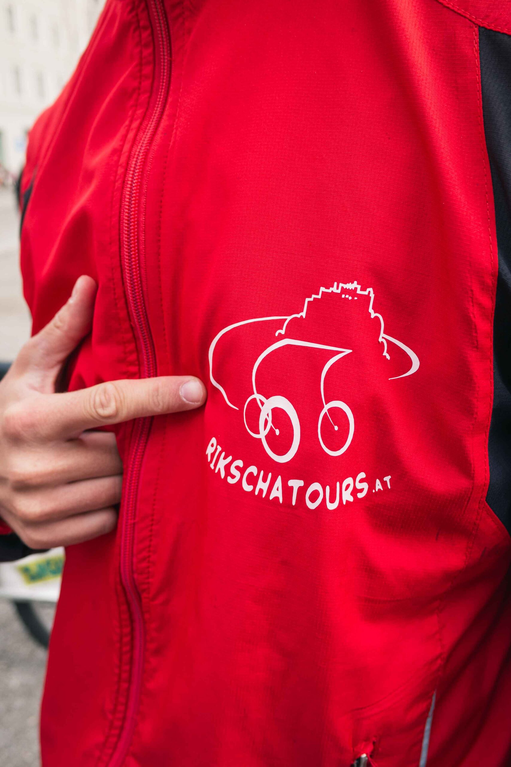 Eine Person zeigt auf ein weißes "Rikscha Tours"-Logo, das eine stilisierte Rikscha auf der Brust einer roten Jacke zeigt.