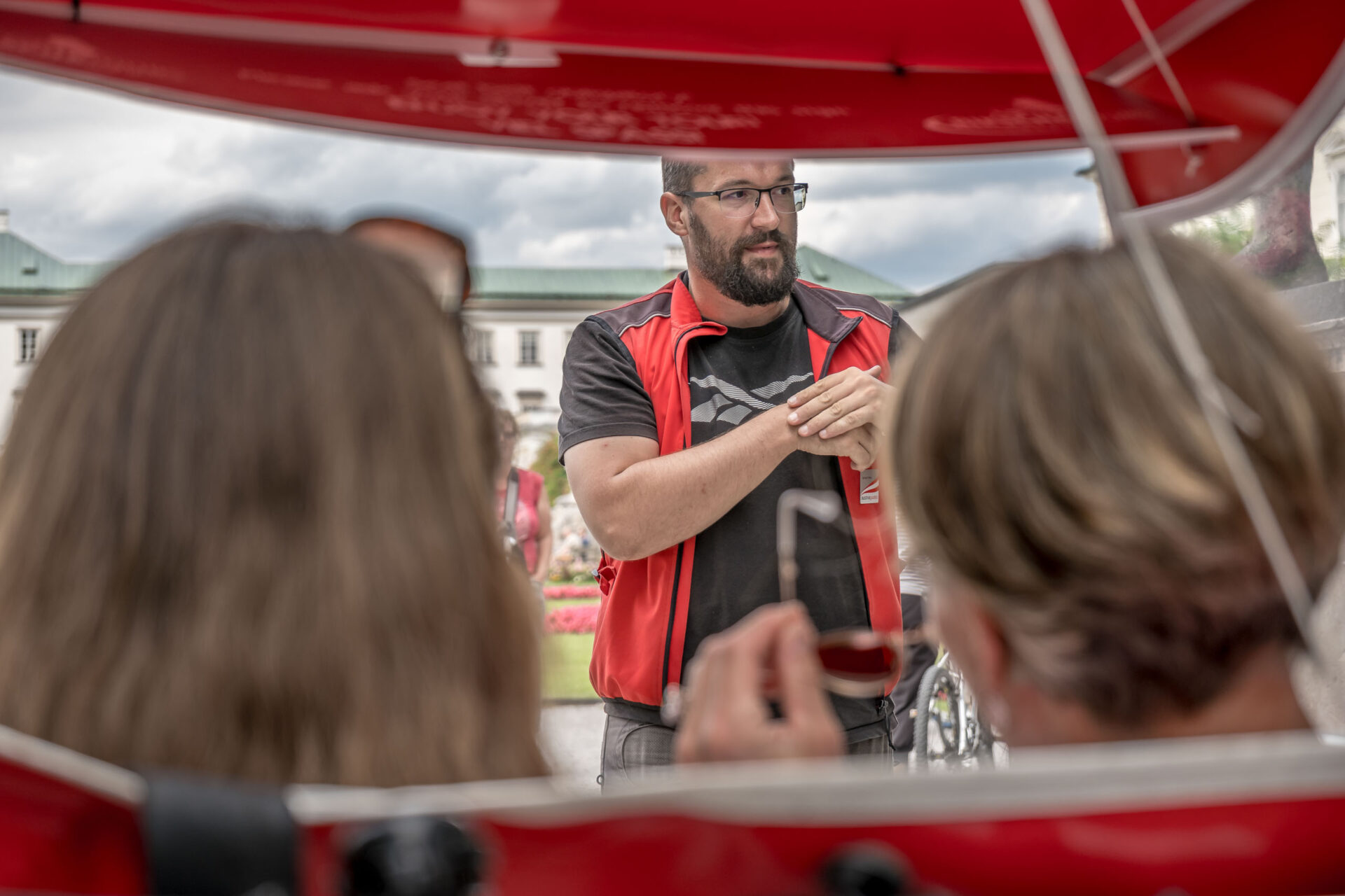 Ein Mann mit roter Weste spricht mit zwei Personen, die vor ihm im Freien sitzen, wobei im Hintergrund Gebäude und Blumen zu sehen sind.