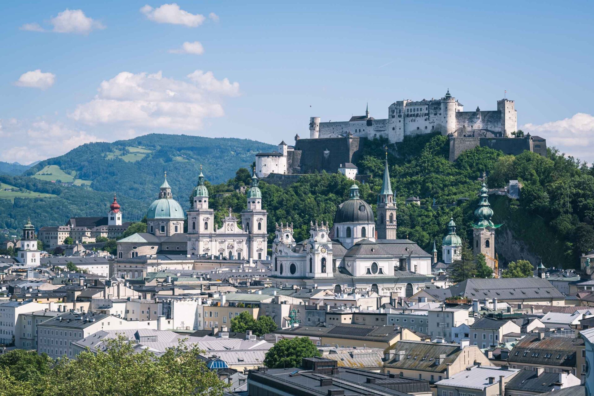 Ein Panoramablick auf Salzburg, Österreich, mit historischen Gebäuden mit Kuppeln und Türmen und der Festung Hohensalzburg auf einem Hügel im Hintergrund unter blauem Himmel.