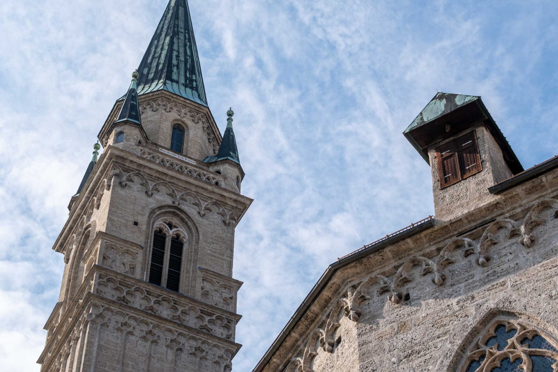 Ein steinerner Kirchturm mit grüner Spitze und Rundbogenfenstern steht neben einem Gebäude mit einem kleinen Holzfenster vor einem teilweise bewölkten Himmel.