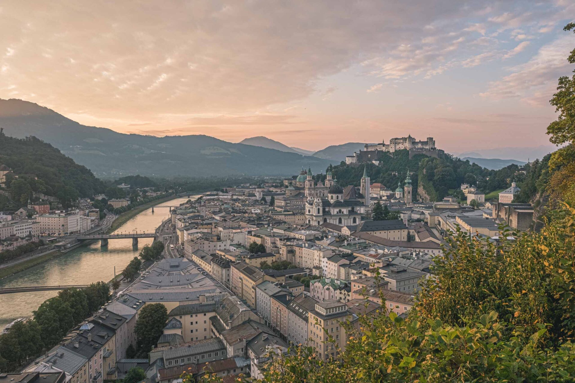 Ein Panoramablick auf Salzburg, Österreich, bei Sonnenuntergang, der die Salzach, historische Gebäude und die Festung Hohensalzburg auf einem Hügel im Hintergrund zeigt.