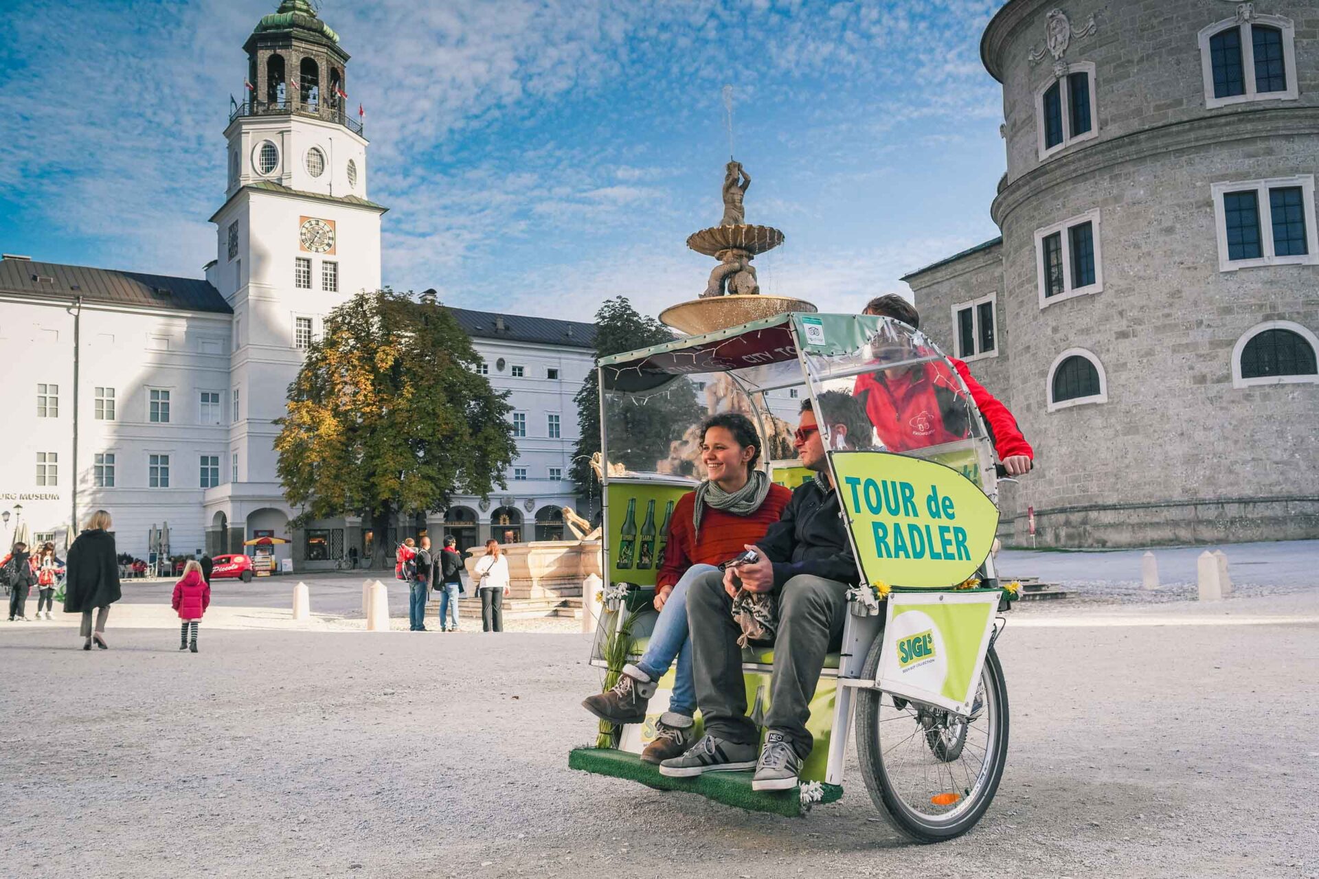 Ein Fahrradtaxi mit der Aufschrift "Tour de Radler" befördert zwei Fahrgäste über einen historischen europäischen Platz mit alten Gebäuden, einem Brunnen und Spaziergängern im Hintergrund.
