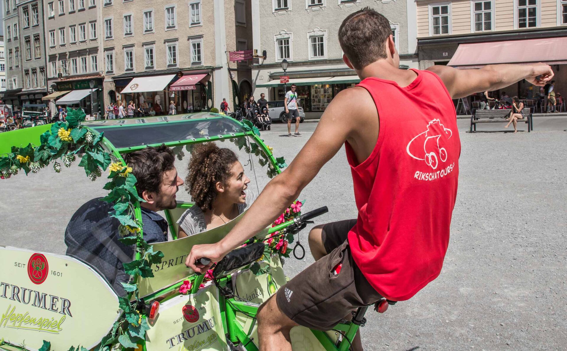 Ein Mann in einem roten Tank-Top tritt in die Pedale eines geschmückten Fahrrads mit zwei Fahrgästen und zeigt auf einem europäischen Stadtplatz auf etwas.