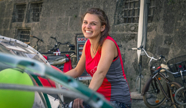Eine junge Frau in einem rot-grauen ärmellosen Hemd lächelt, während sie im Freien neben Fahrrädern und einer Steinmauer steht.