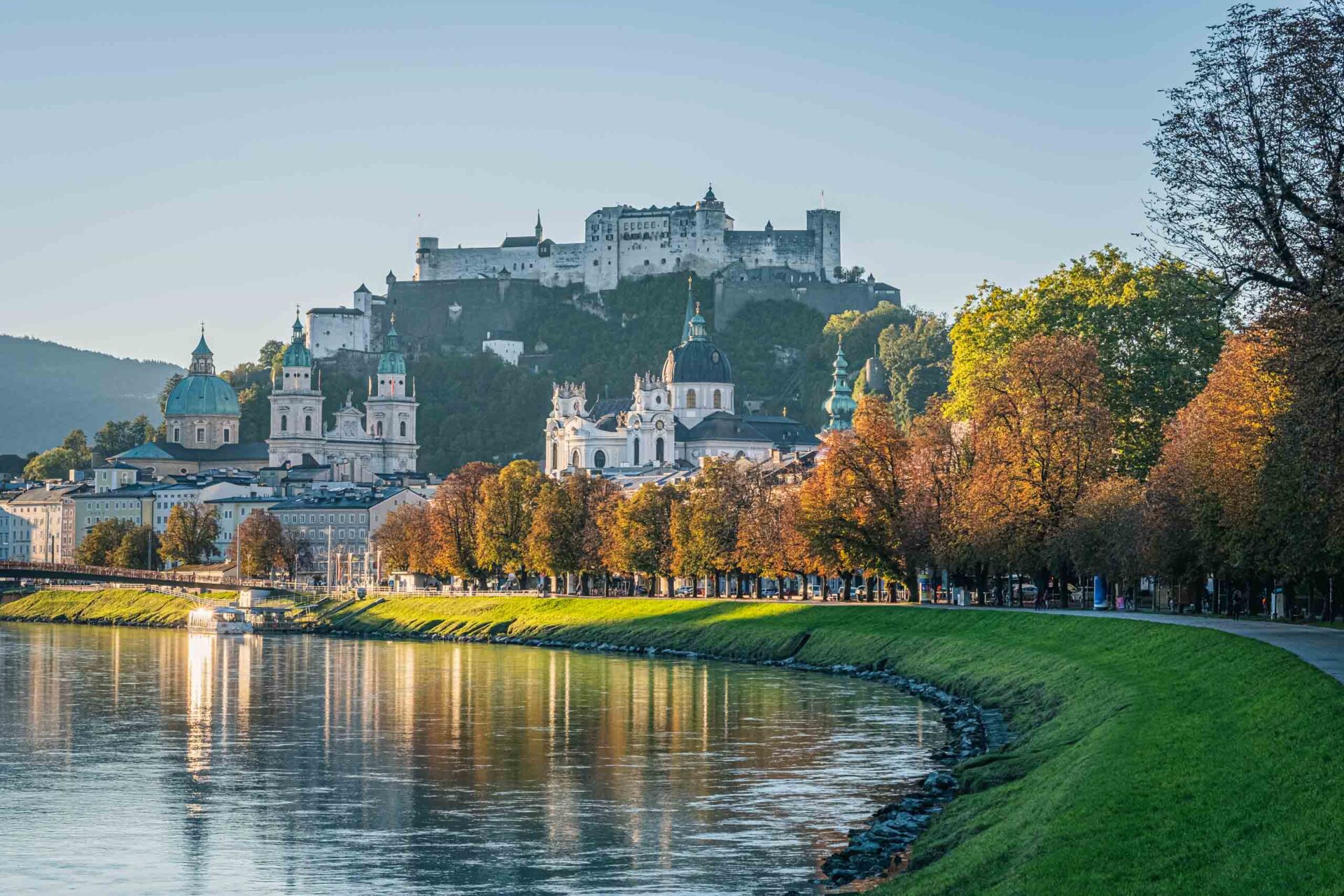 Ein Blick auf das Flussufer von Salzburg, Österreich, mit historischen Gebäuden und der Festung Hohensalzburg auf einem Hügel im Hintergrund. Bäume säumen das Flussufer.