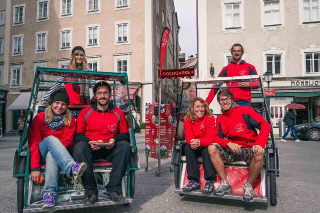 Sechs Personen in passenden roten Hemden sitzen und stehen hinter zwei Rikschas auf einer Stadtstraße mit alten Gebäuden im Hintergrund.