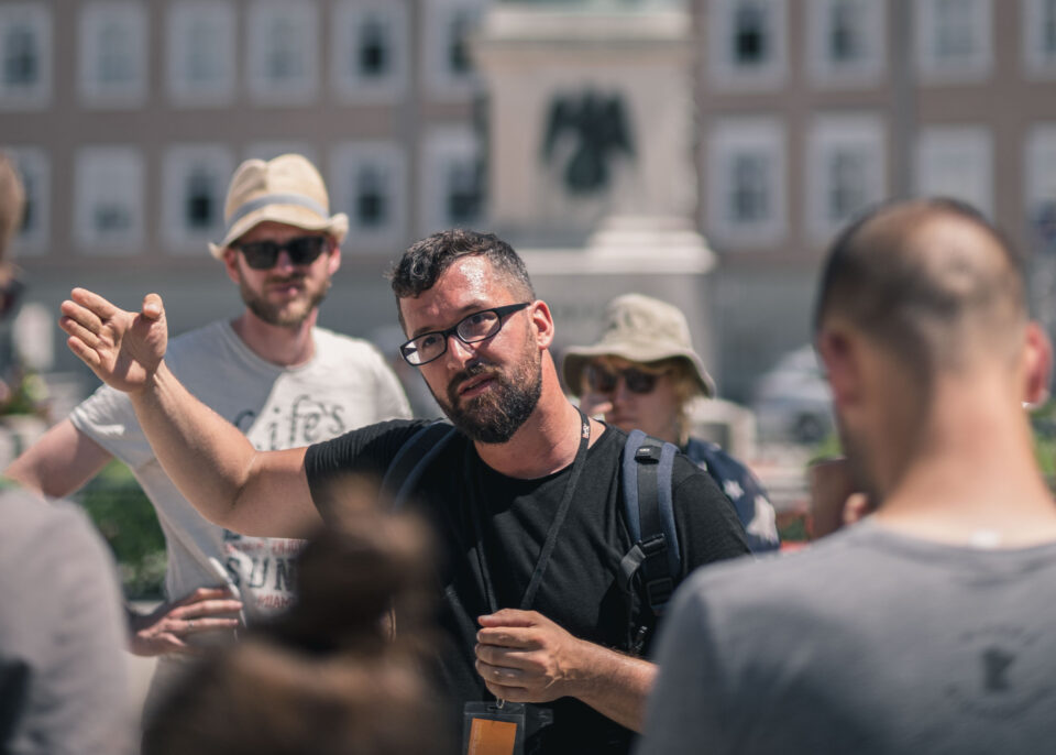 Ein Mann mit Brille und Rucksack gestikuliert, während er zu einer Gruppe von Menschen im Freien spricht, mit Gebäuden im Hintergrund.