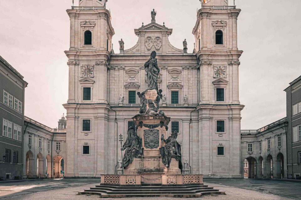 Eine große Steinstatue steht in der Mitte eines Hofes vor einer kunstvollen, historischen Kathedrale mit Zwillingstürmen und Bogenfenstern.