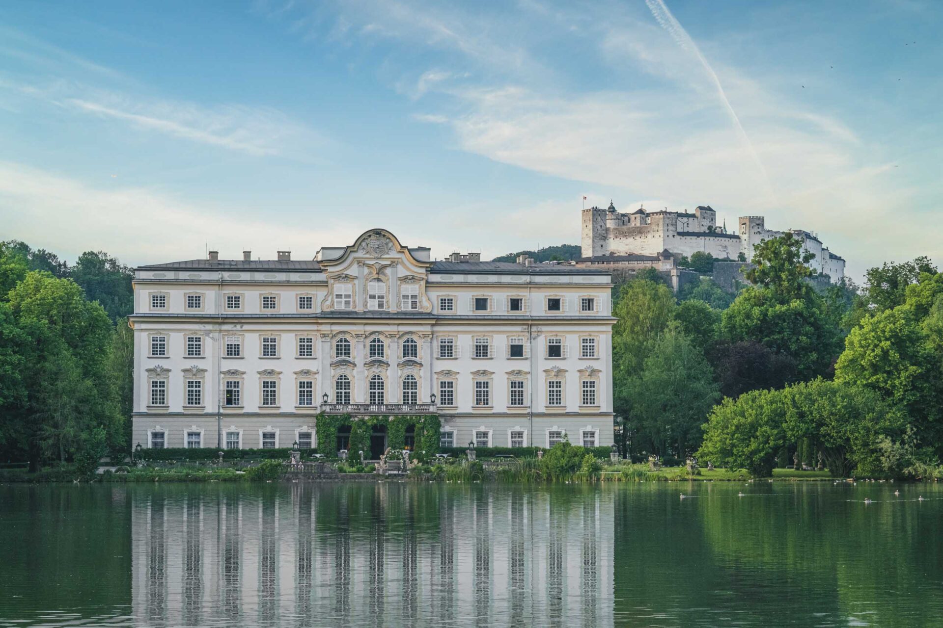 Ein großer weißer Palast spiegelt sich in einem ruhigen, von Bäumen umgebenen See, während im Hintergrund eine Festung auf einem Hügel unter blauem Himmel zu sehen ist.