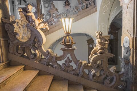 Verschnörkelte Steintreppe mit dekorativem Geländer, zwei Cherub-Statuen und einer zentralen Vintage-Lampe in einem kunstvoll gestalteten Innenraum.