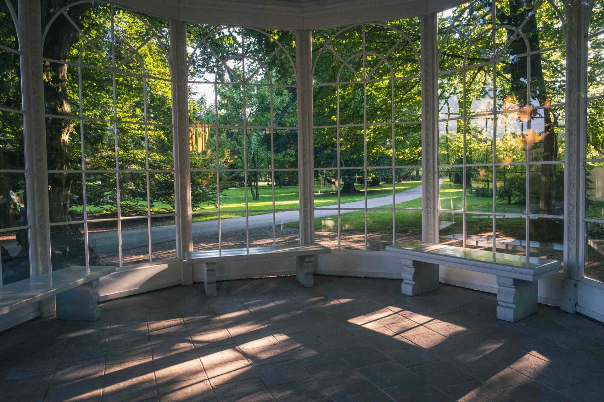Blick auf einen grünen Park durch große Glasfenster aus dem Inneren eines Pavillons, wobei das Sonnenlicht Schatten auf den gefliesten Boden und die Bänke wirft.